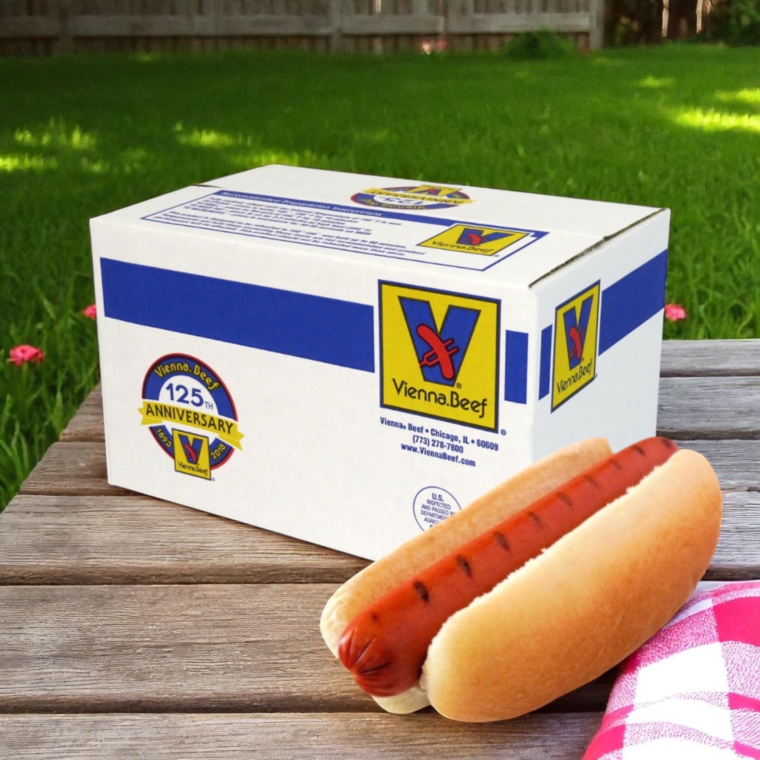 A Vienna Beef Halal Skinless Beef Frank 6” 5:1 in a bun sits on a picnic table beside a Vienna Beef 125th anniversary box, with grass and a fence in the background.