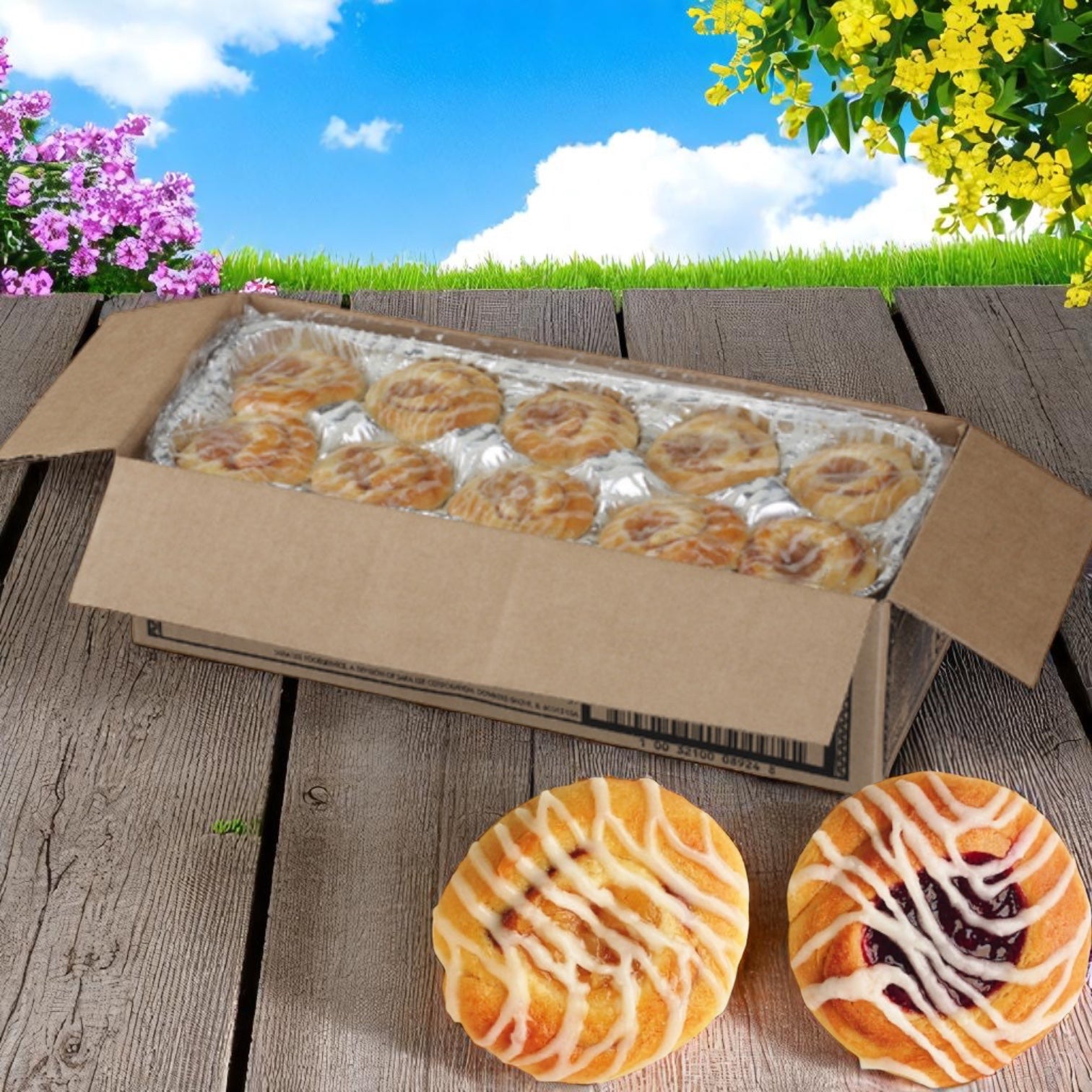An open box of Chef Pierre Danish Assorted Mini 1.25 OZ pastries sits outdoors on wood, with two flaky, iced pastries in front; grass, flowers, and blue sky add a fresh backdrop to the Chef Pierre snack display.