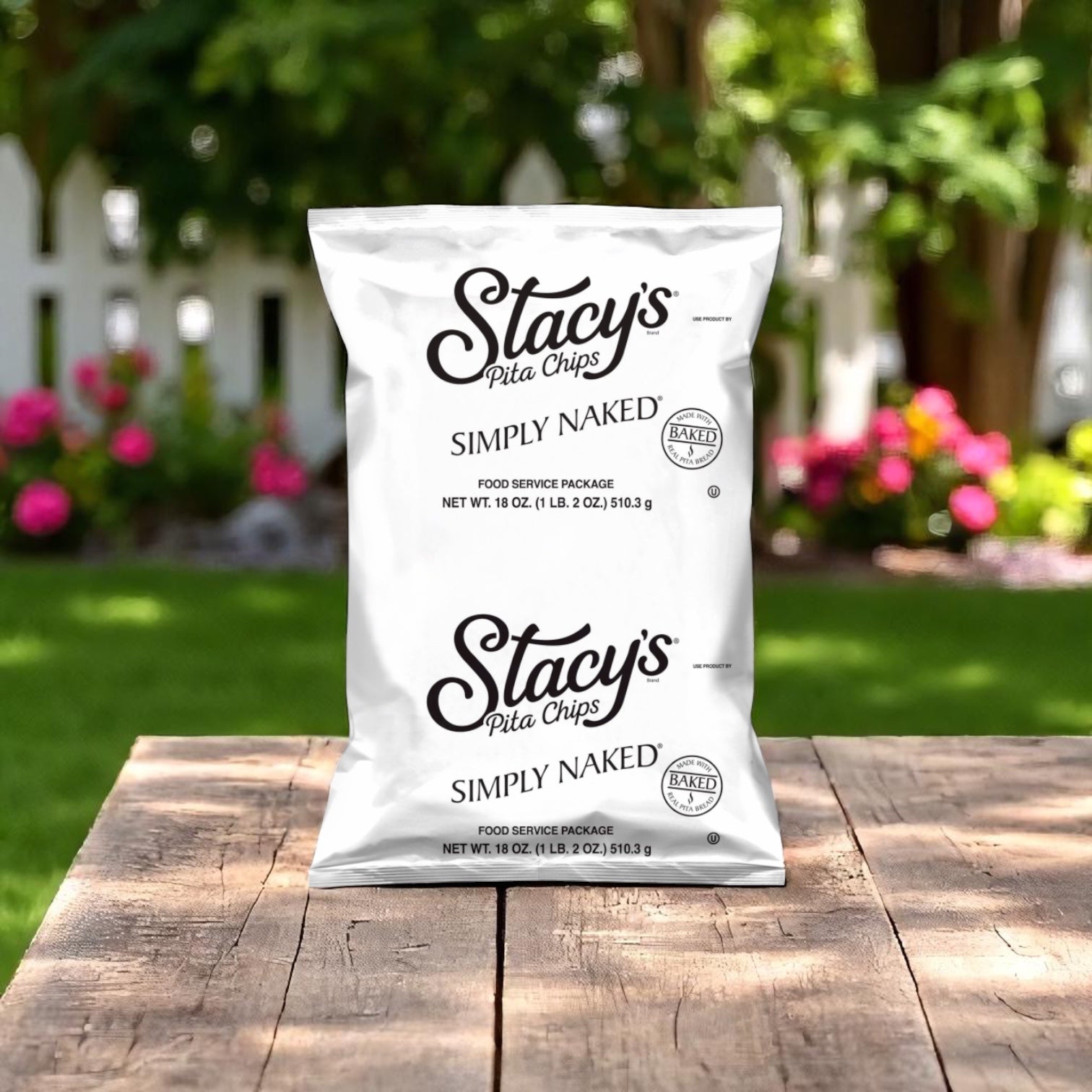 A 16 oz bag of Easy Lunches Stacys Simply Simply Naked Pita Chips (6 count) is on a wooden table outdoors, with a white fence and blooming flowers in the background.