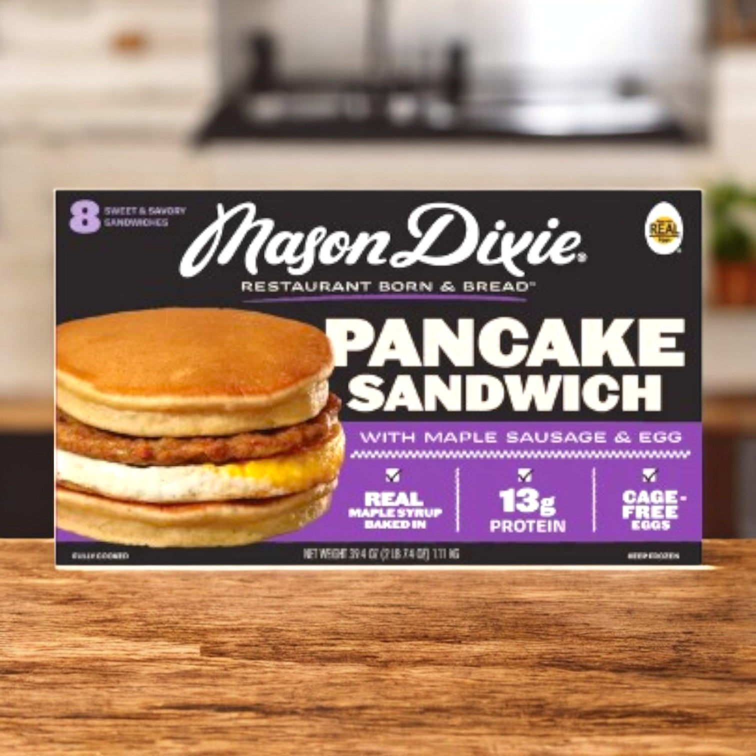 An 8-count case of Easy Lunches Mason Dixie Pancake Sandwich sits on a kitchen counter. The breakfast sandwich packaging highlights 13g protein, real maple syrup, and cage-free eggs.