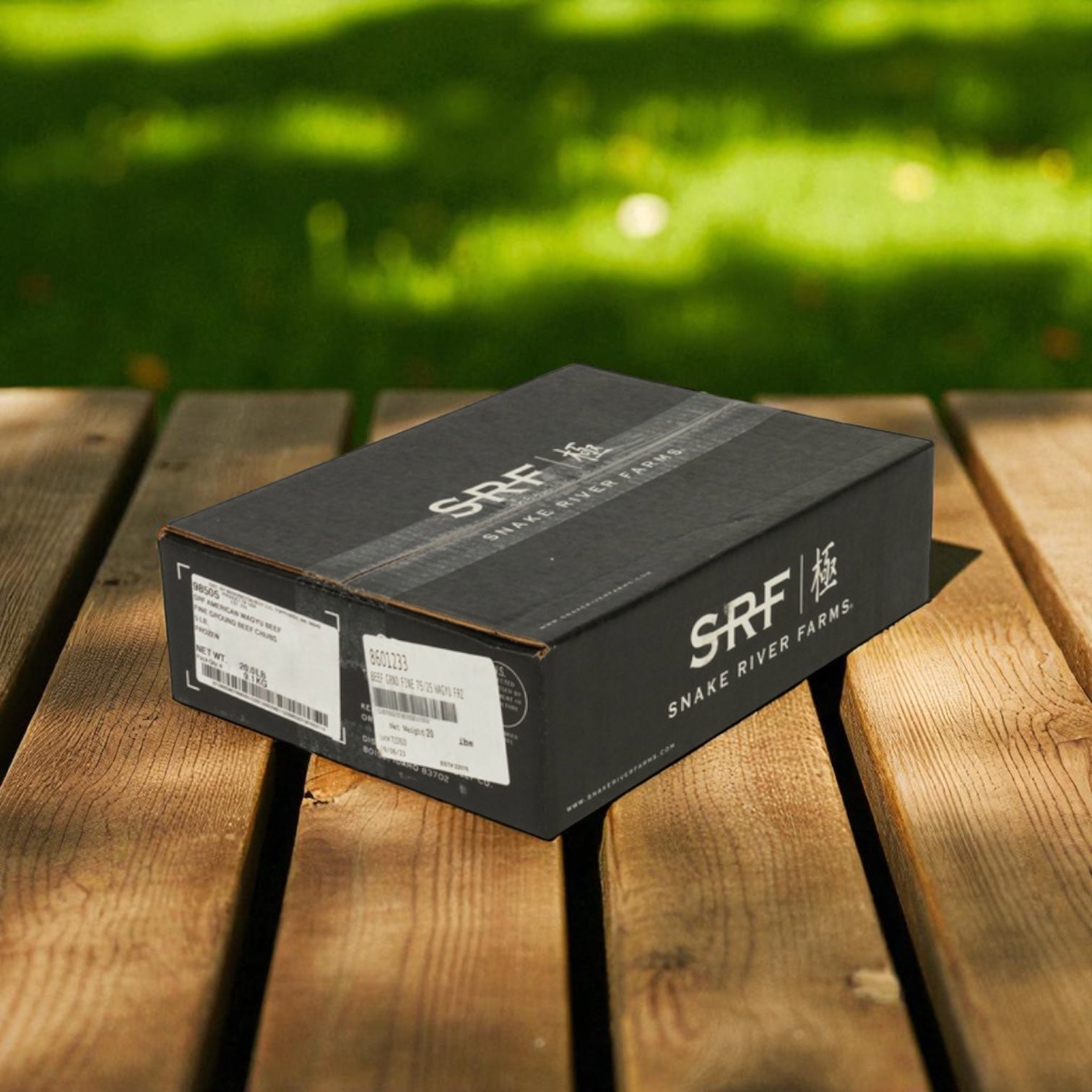A package of Snake River Farms American Style Wagyu Beef Ground 75% (4 Count, 5 lb) rests on a wooden surface outdoors, sunlight and grass visible in the background.