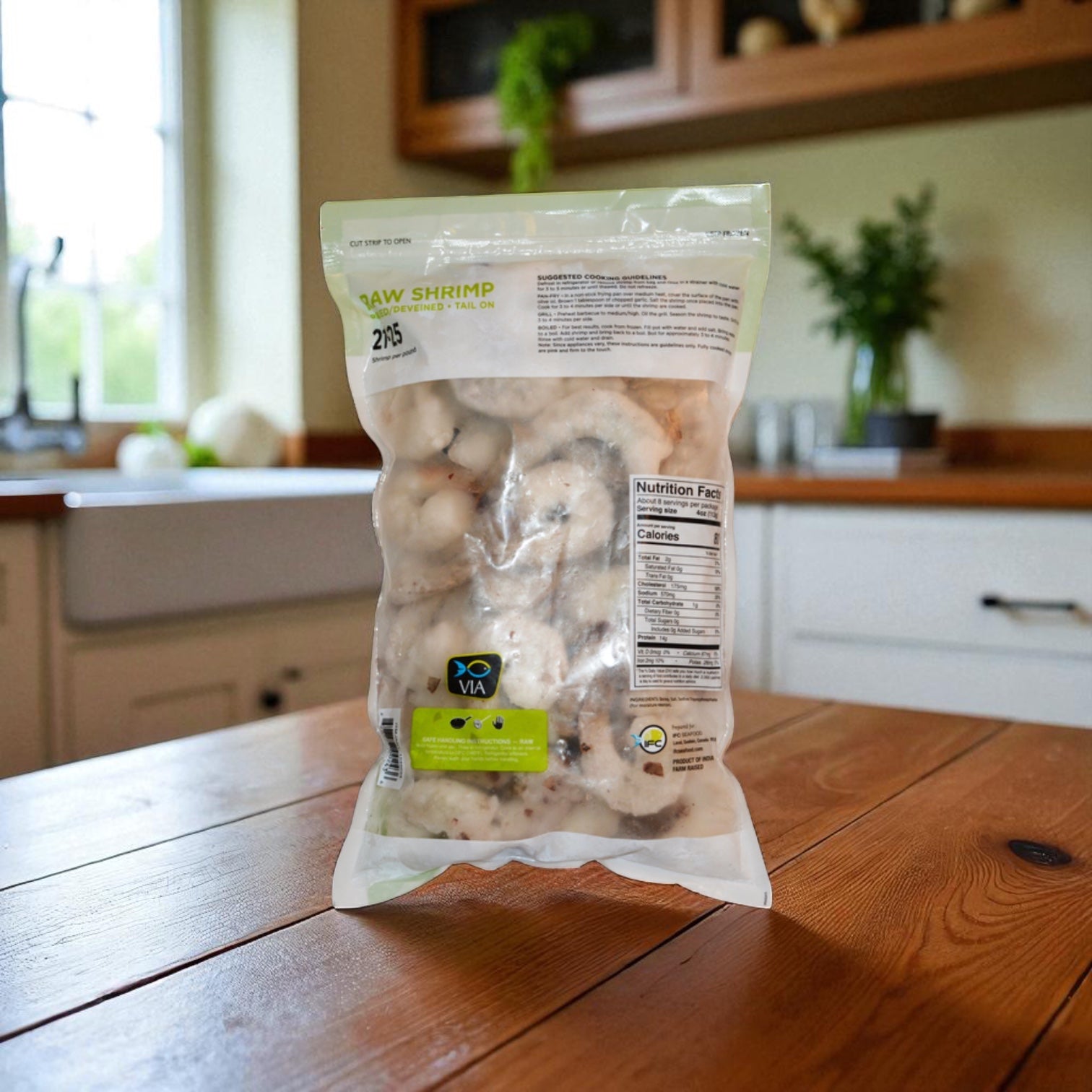 A sealed bag of Easy Lunches VIA Farm-Raised Raw Shrimp, Tail-On, Peeled, Deveined, 21-25 ct, 2 lbs rests on a wooden kitchen table with a blurred background of a sink and plants.