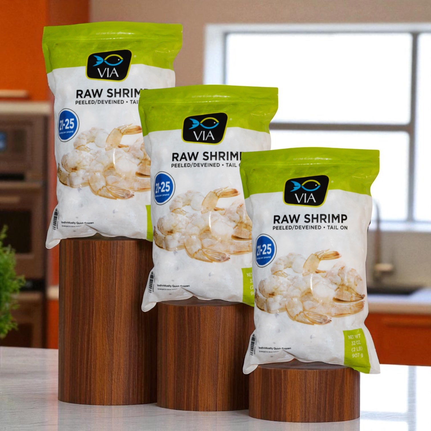 Three packs of Via Farms VIA Farm-Raised Raw Shrimp, Tail-On, Peeled, Deveined, 21-25 ct, 2 lbs each are displayed on wooden stands in a kitchen.