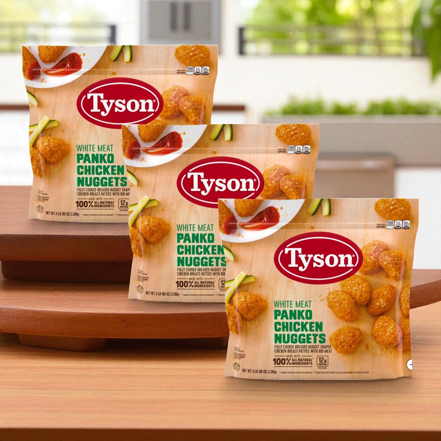 Three bags of Tyson Panko Chicken Nuggets, 5 lbs each (3 count), are arranged on a wooden surface with a blurred indoor background.