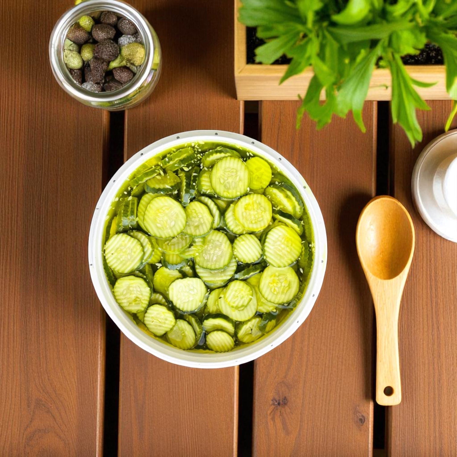 A bowl of sliced pickles in brine sits on a wooden table beside a spoon, jar of olives, and potted plant—just like the tasty Chipico Kosher Dill Chips from Vienna Beef’s famous 2 Gallon Jar with 450 chips.