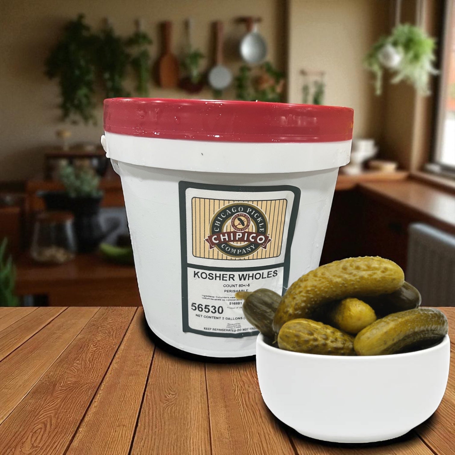 A white plastic Chipico Kosher Wholes 2 Gallon Container by Vienna Beef sits on a table beside a bowl of kosher dill pickles, with a kitchen in the background.