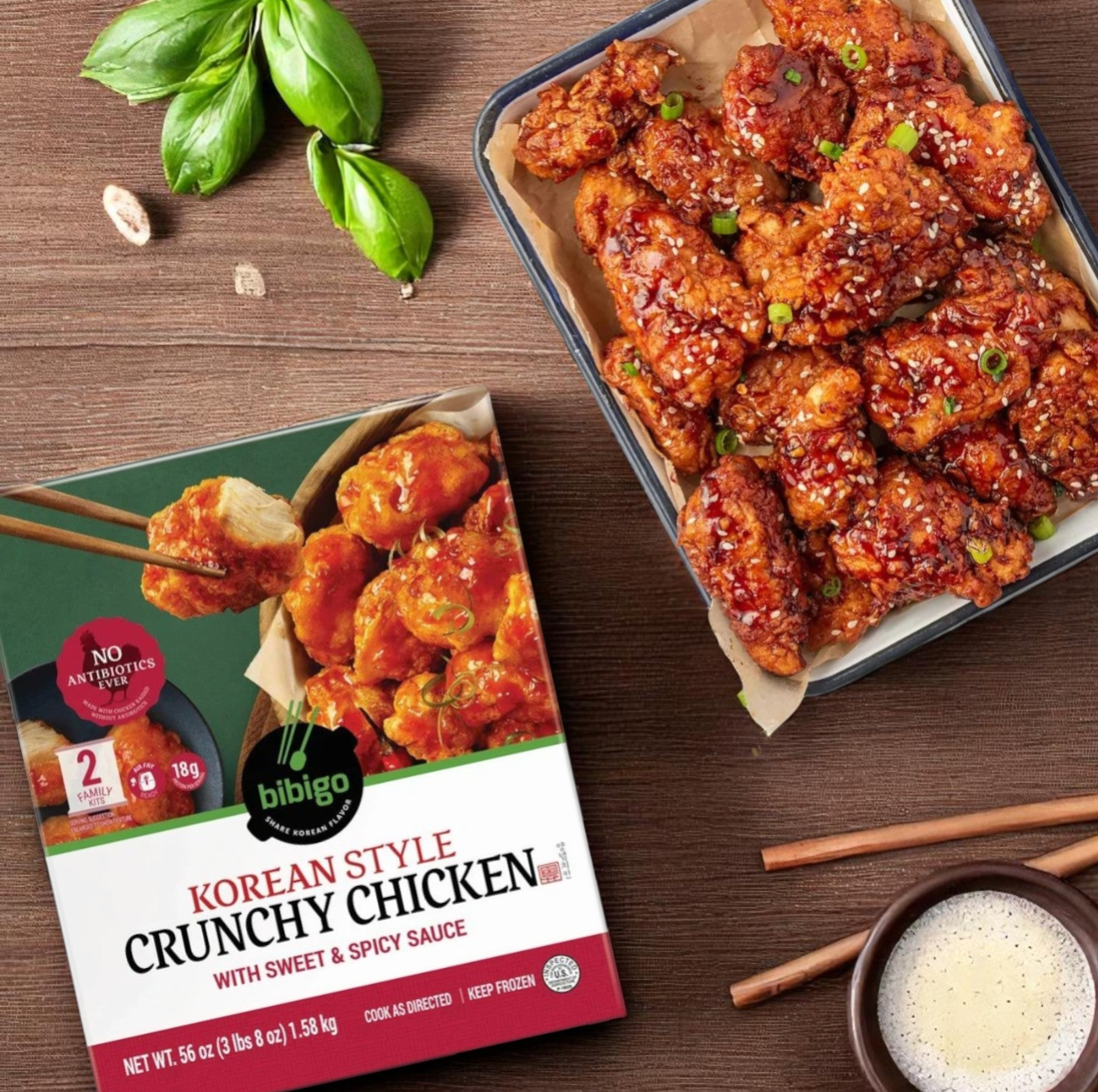 A 56 oz bag of Easy Lunches Bibigo Korean Style Crunchy Chicken with Sweet & Spicy Sauce sits beside a tray of garnished chicken pieces, chopsticks, basil leaves, and sauce.