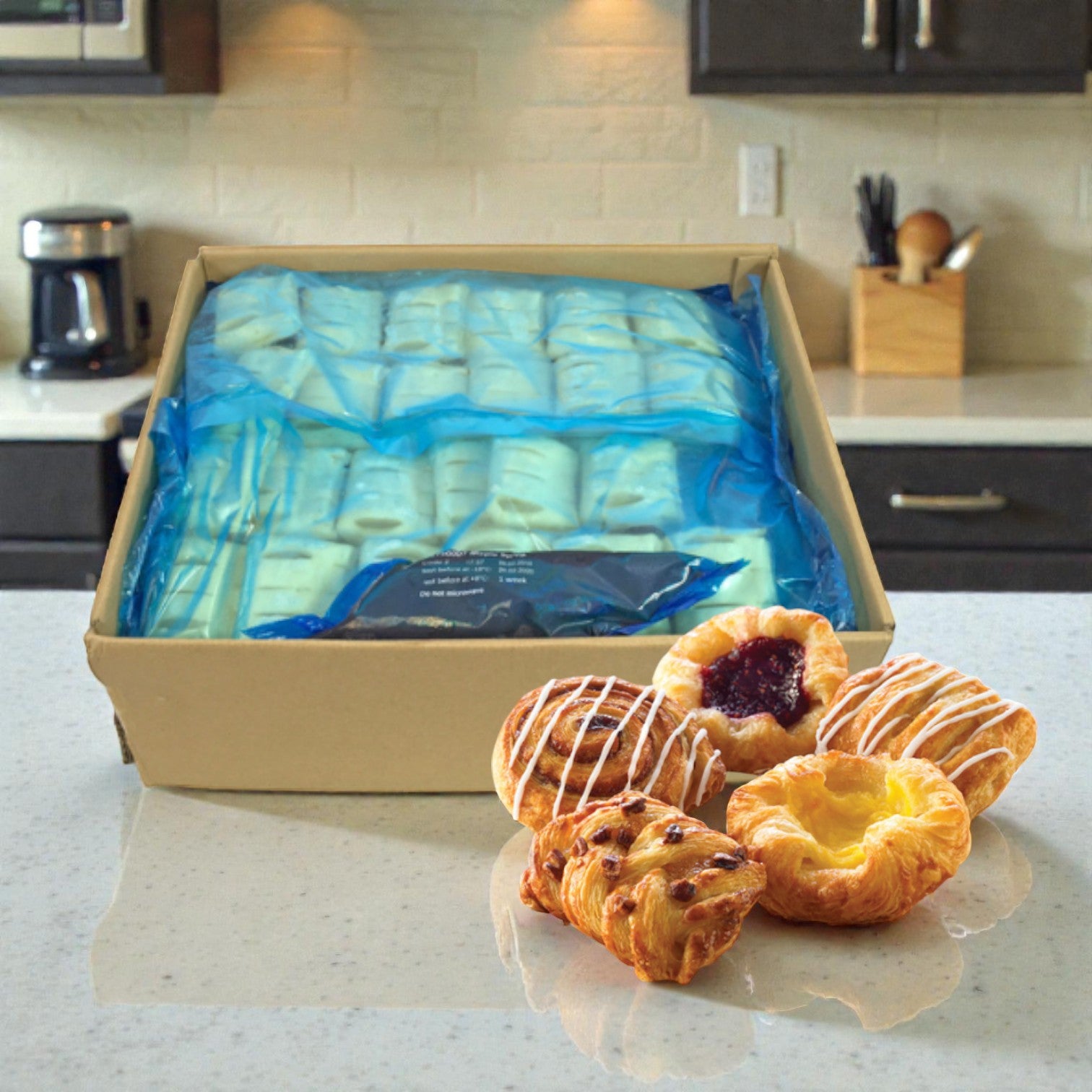 A Schulstad USA Inc Mini Danish Pastry Selection (120 count, 1.5 oz each) in blue packaging sits on a kitchen counter beside assorted pastries, evoking the taste of authentic European Danish treats.