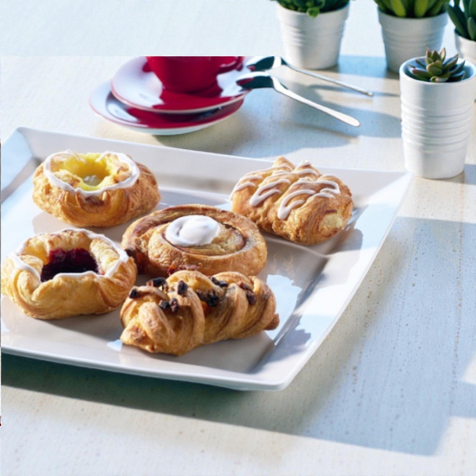 A Schulstad USA Inc Mini Danish Pastry Selection (120 count, 1.5 oz each) is served on a white square plate atop a light table, with a red cup, saucer, utensils, and potted plants in the background.