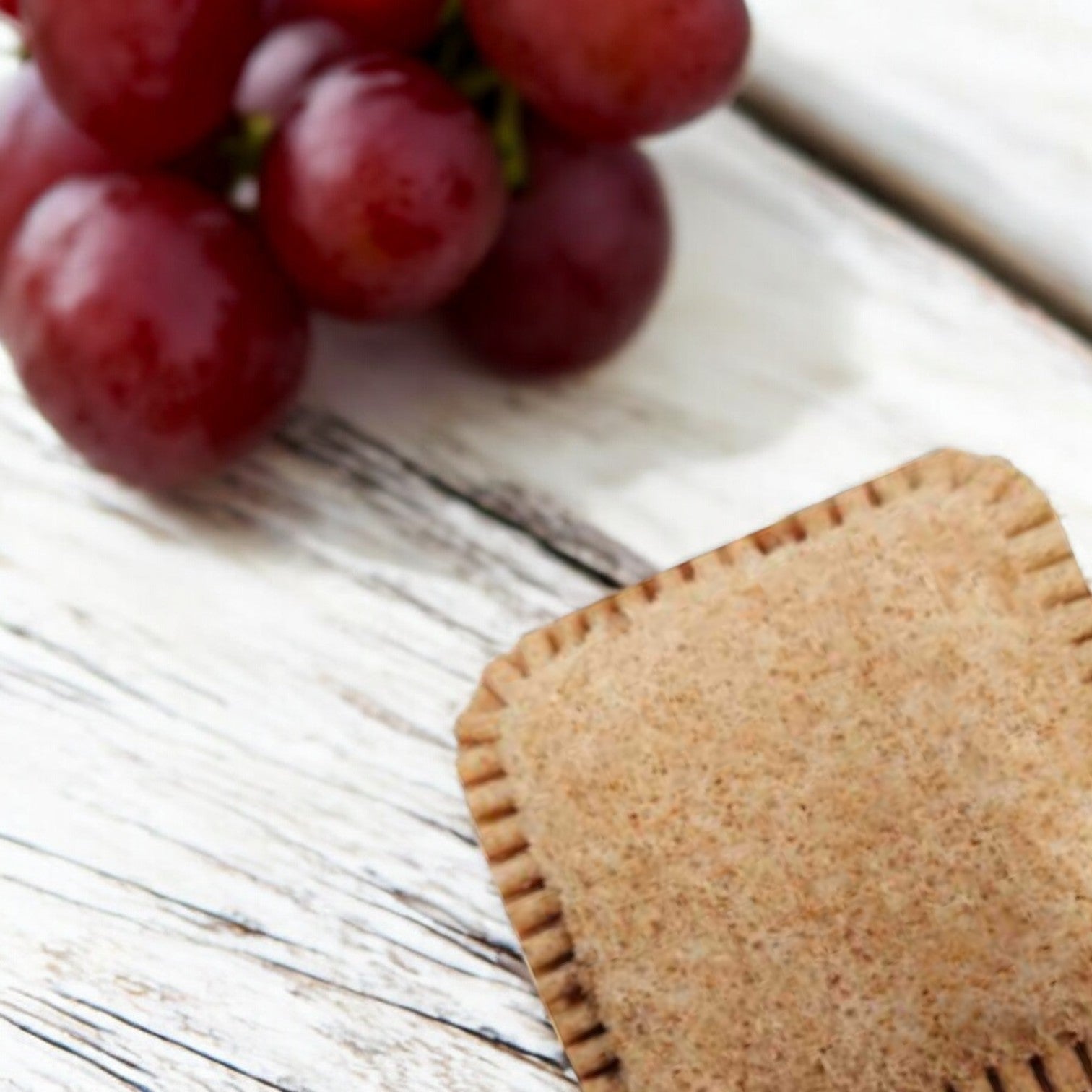 A group of red grapes and a square pastry on a white wooden surface, perfect companions to nut-free snacks like Sunbutter Jammies Sandwich Sunflower Butter & Grape Jelly from Sunbutter.