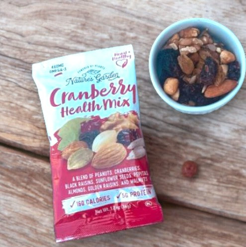 A packet of Nature's Garden Cranberry Health Trail Mix, a 1.2 oz serving labeled as healthy and lightly salted, sits beside a bowl of mixed nuts and dried fruits on a wooden surface, offering heart-healthy snacks perfect for any time.