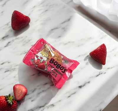 A Bobo's Strawberry Stuff'd Oat Bite (1.3 oz) in its pink packaging sits on a marble surface, surrounded by three whole strawberries and one sliced strawberry half, emphasizing its gluten-free, vegan goodness.