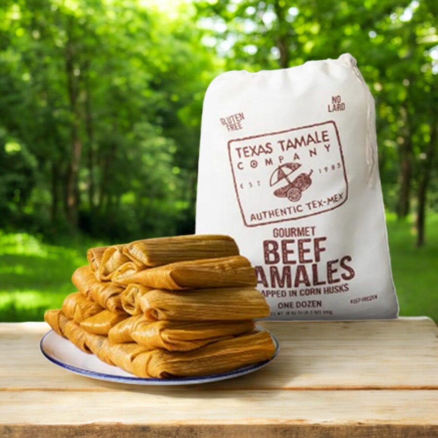 An 18 oz bag of Texas Tamale Company Beef Tamales sits on a table alongside gluten-free tamales wrapped in corn husks. Set against a lush forest backdrop, they exude authentic Tex-Mex flavor.