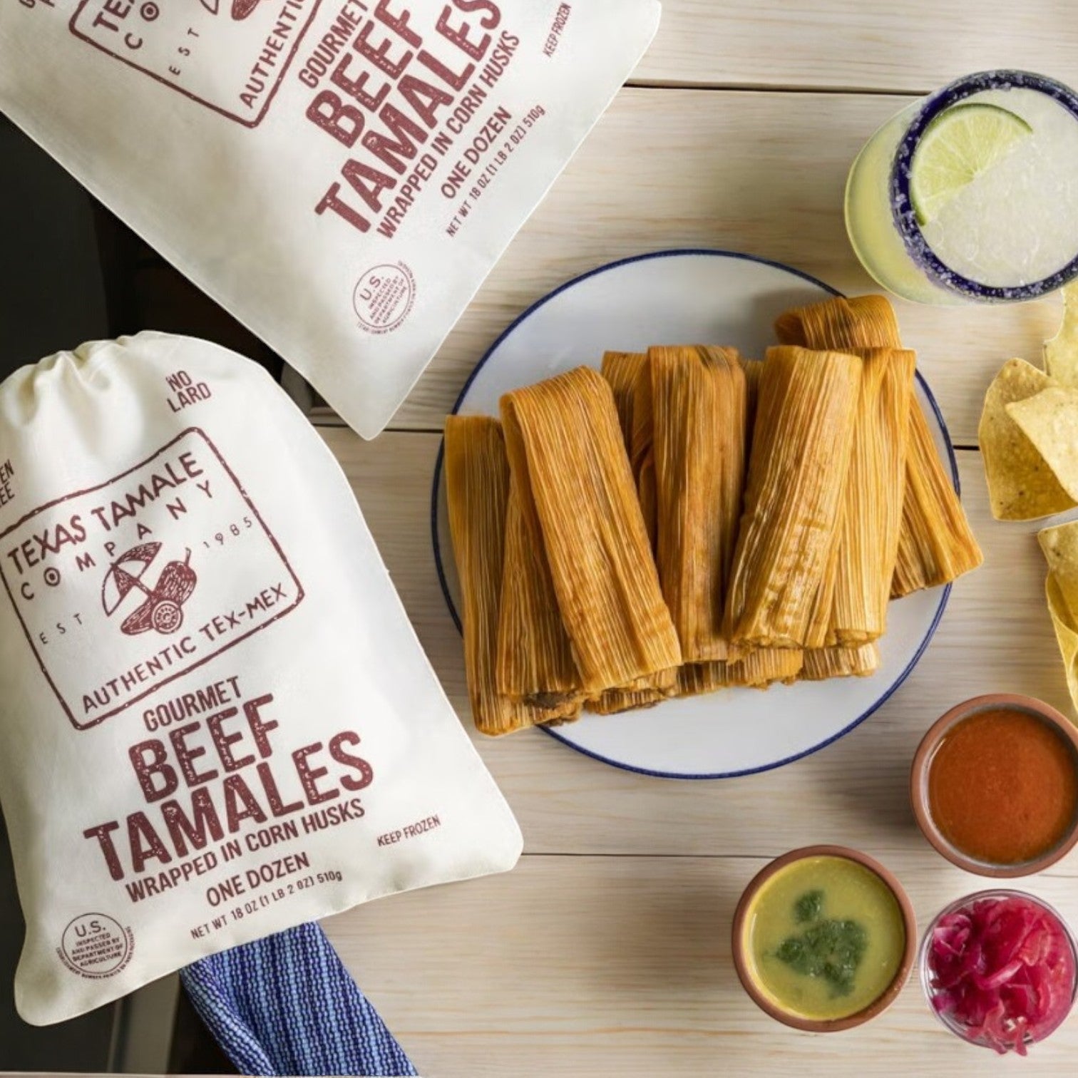 Texas Tamale Company's gluten-free Beef Tamales, 18 oz bag, offer rich Tex-Mex flavor. They sit beside a feast of unwrapped tamales, tortilla chips, red and green sauces, and two ice-cold drinks—guaranteed to elevate your dining experience.