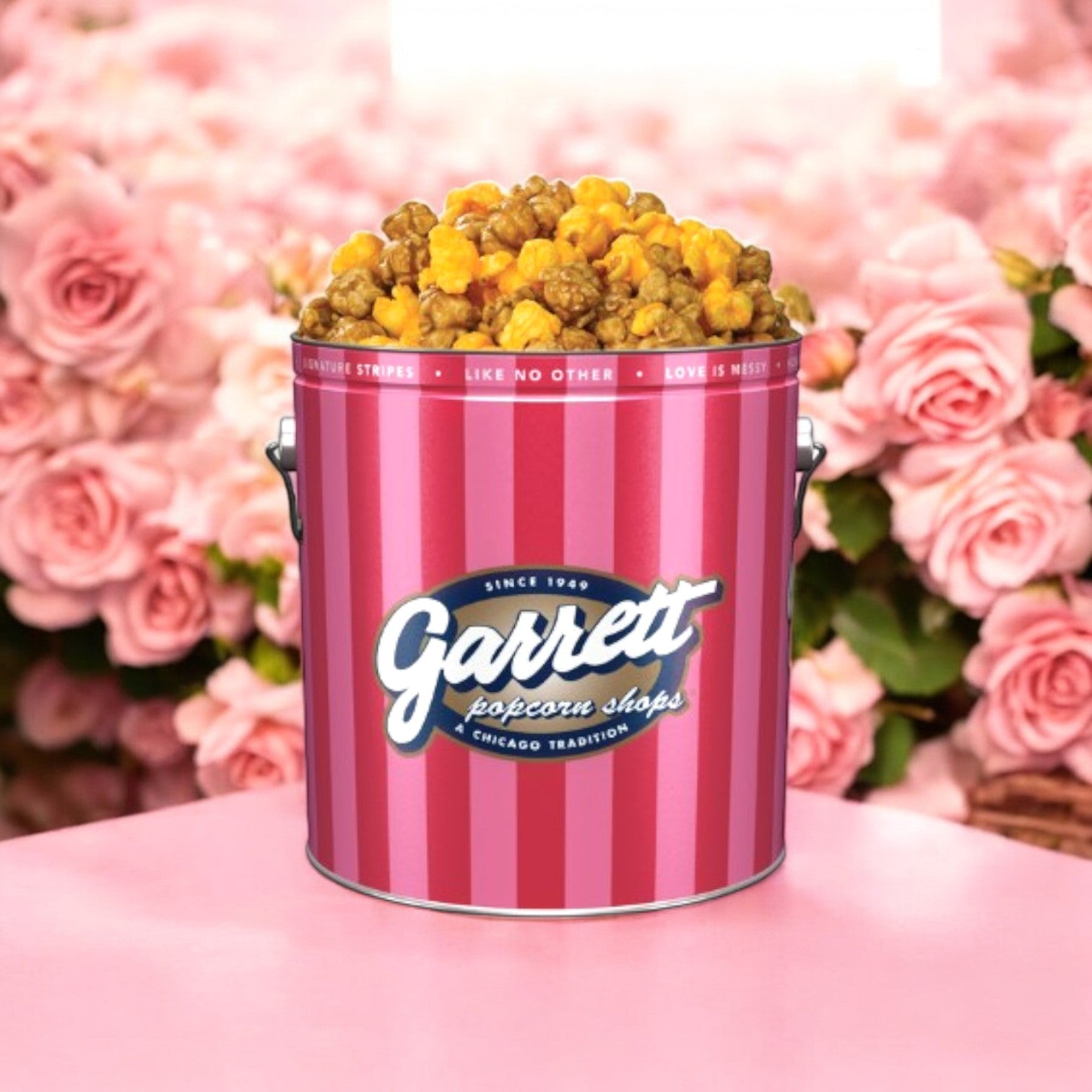 A tin of The Garrett Mix and Fannie May Pixel Combo! by Easy Lunches, offering a sweet and savory caramel cheese popcorn treat, sits on a table. Its pink and red striped design beautifully complements the backdrop of pink roses.