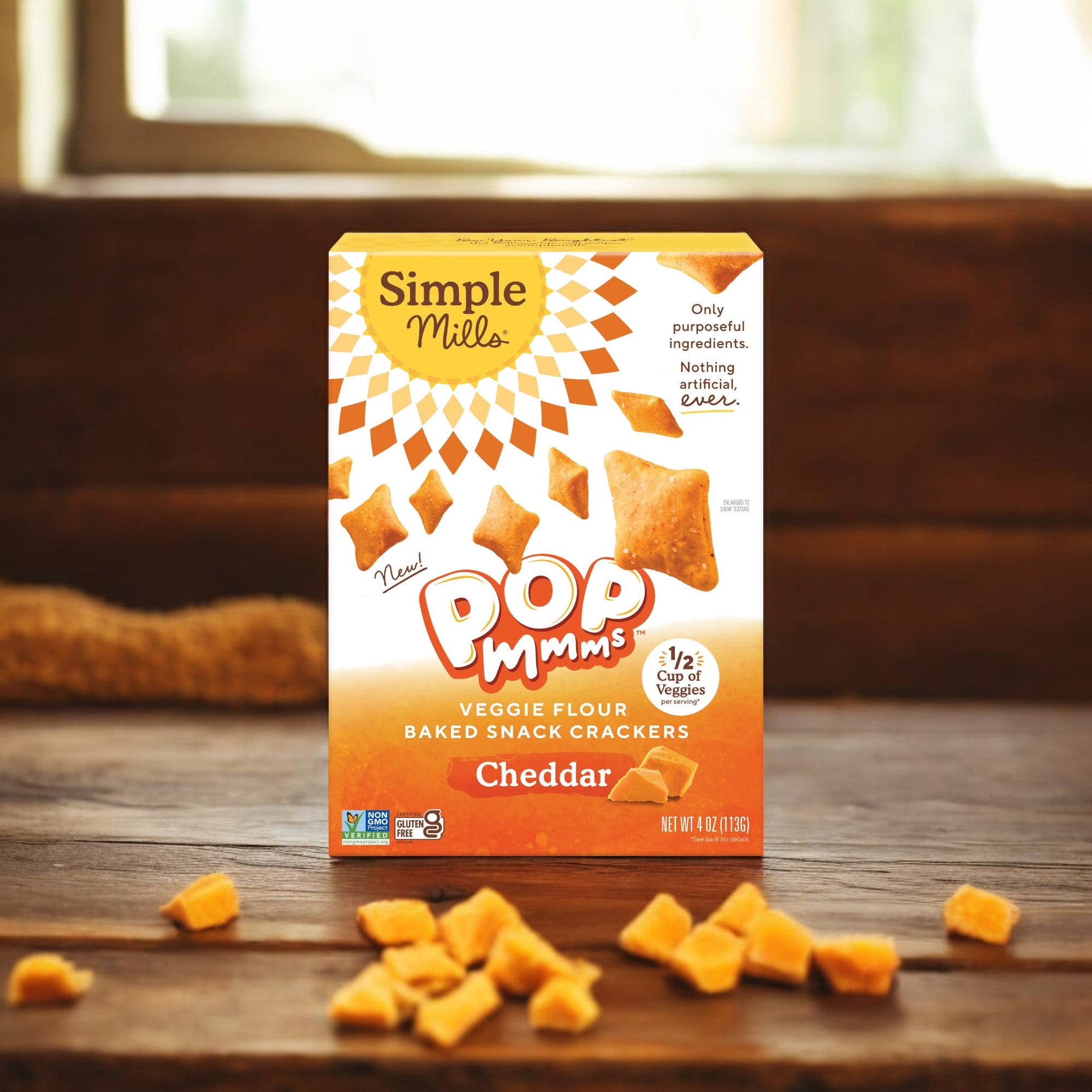 A box of Simple Mills Cheddar Pop Mmms, Gluten Free Baked Snack Crackers (4 oz) sits on a wooden surface, surrounded by some scattered butternut squash crackers. Perfect for healthy snacking at any time of day, these veggie-packed treats are an ideal choice.