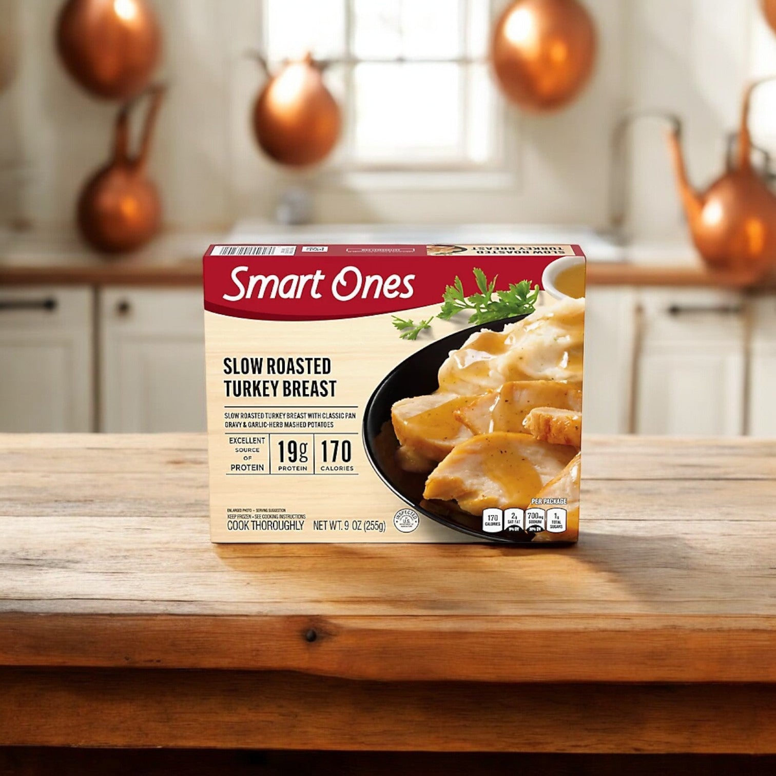 A Smart Ones® box of Weight Watchers® Slow Roasted Turkey Breast with Gravy & Garlic-Herb Mashed Potatoes, 9 oz. - 12 Count, rests on a wooden countertop in a kitchen setting with white cabinets and copper utensils in the background.