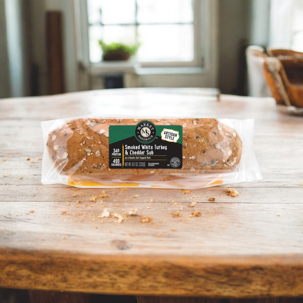 An Easy Lunches Smoked Turkey & Cheddar Sub Sandwich on an artisan sub roll sits on a wooden table, with a blurred interior background.