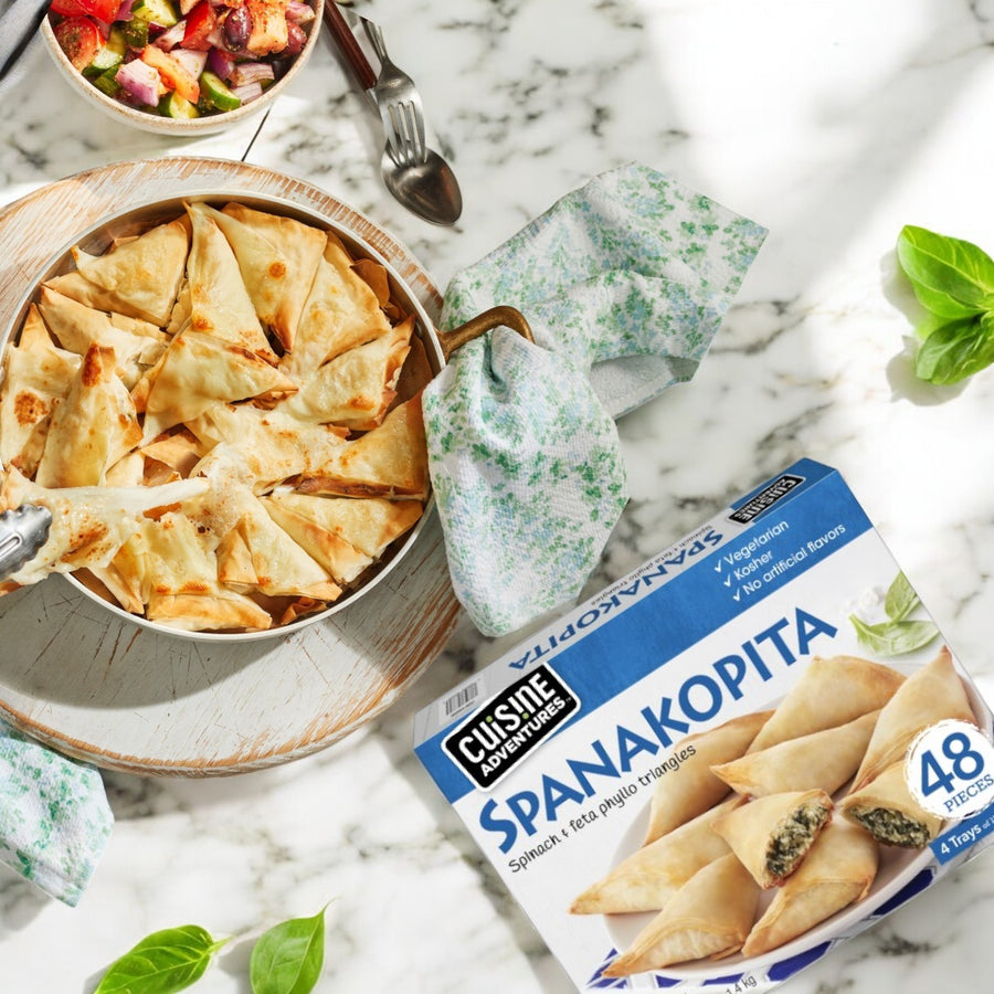 A bowl of Cuisine Adventures Spanakopita Spinach & Feta Phyllo Triangles is next to its 48 oz. package. A vibrant salad and scattered basil leaves enhance the marble surface.