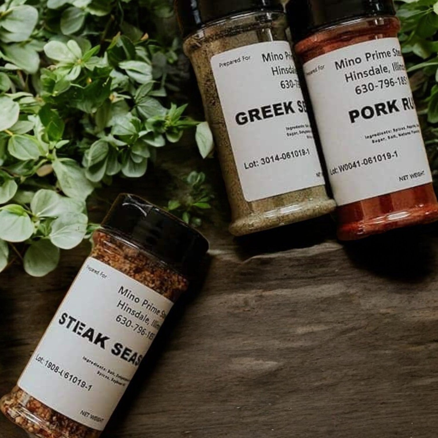 Three spice bottles labeled Greek, Pork Rub, and Mino Prime Steak Seasoning rest on a wooden surface with green leaves nearby, promising steakhouse-quality flavors ideal for crafting those Mino Prime Steak Burgers.