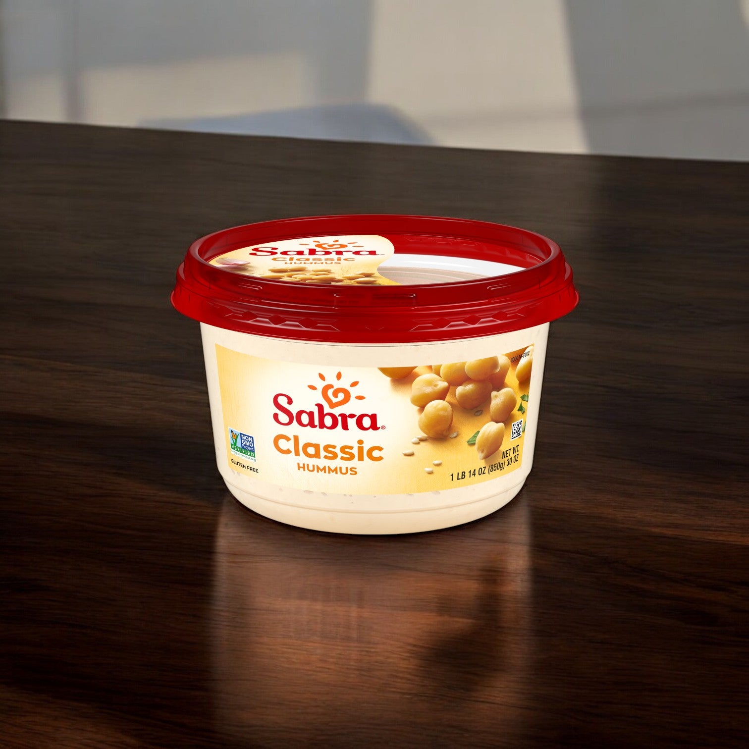 A 30 oz. container of Sabra Classic Hummus Spread with a red lid is placed on a dark wooden surface. The packaging displays images of chickpeas and a label indicating it is non-GMO, promising an authentic flavor and making it a versatile dip for any occasion.