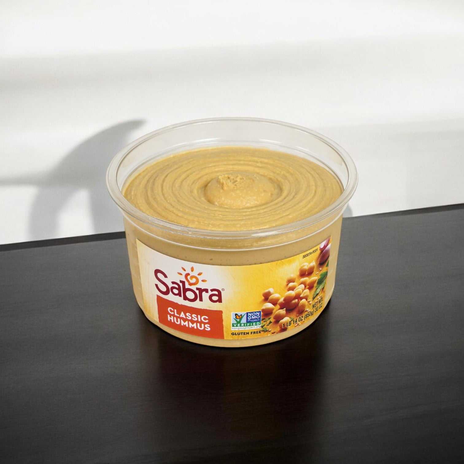 A container of Sabra Classic Hummus Spread, 30 oz. - 1 Count, rests on a dark surface, showcasing its label that proudly indicates it is gluten-free and Non-GMO Project Verified. This versatile dip offers an authentic flavor perfect for any occasion.