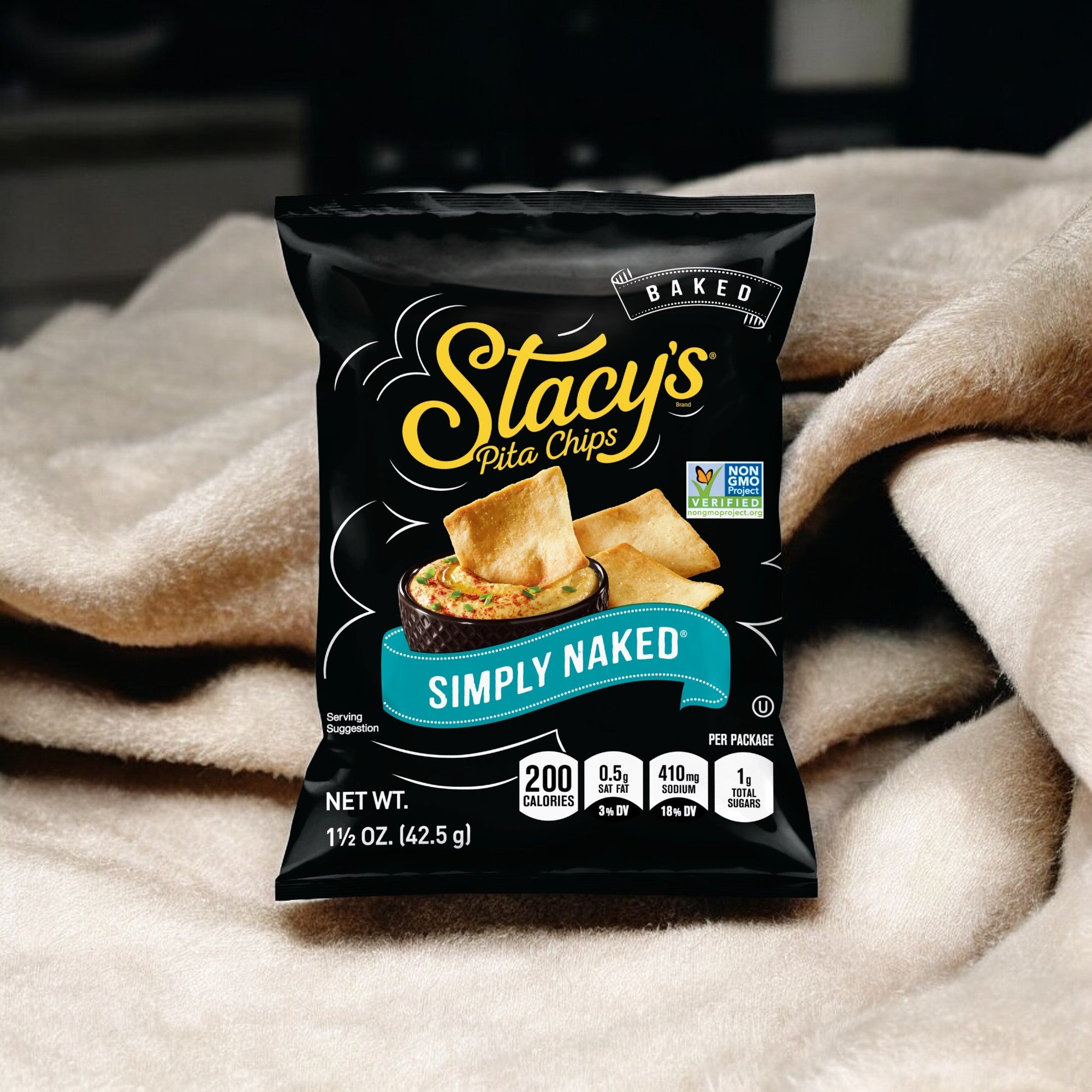 A package of Stacy's Pita Chips Simply Naked 1.5 oz rests on a soft, light-colored blanket. The bag prominently displays product information, including non-GMO certification and nutritional details.