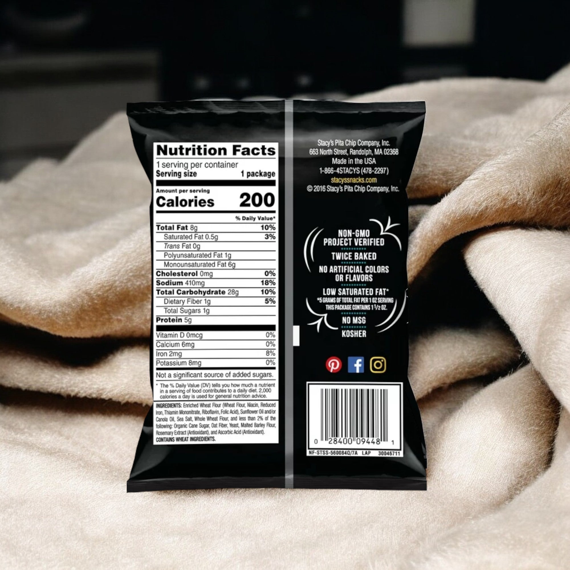 The back of a Stacy's Simply Naked Pita Chips 1.5 oz package displays nutrition facts, dietary information, and logos for non-GMO, gluten-free, and kosher certifications. The sea salted chip package is set on a beige fabric background.