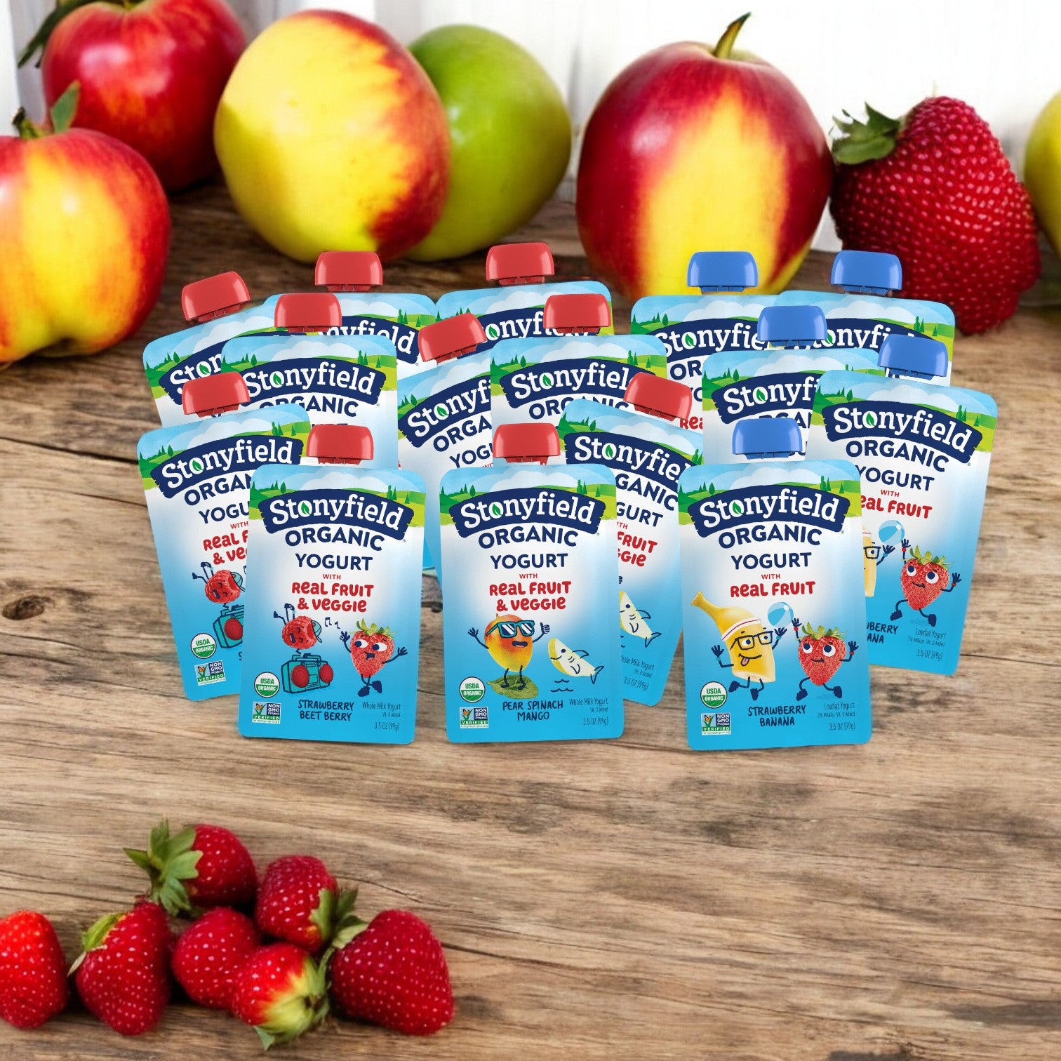 A variety of Stonyfield's Organic kids’ yogurt pouches from the 15 Bundle Pack, featuring three flavors with five of each, are arranged on a wooden surface with apples and strawberries in the background.