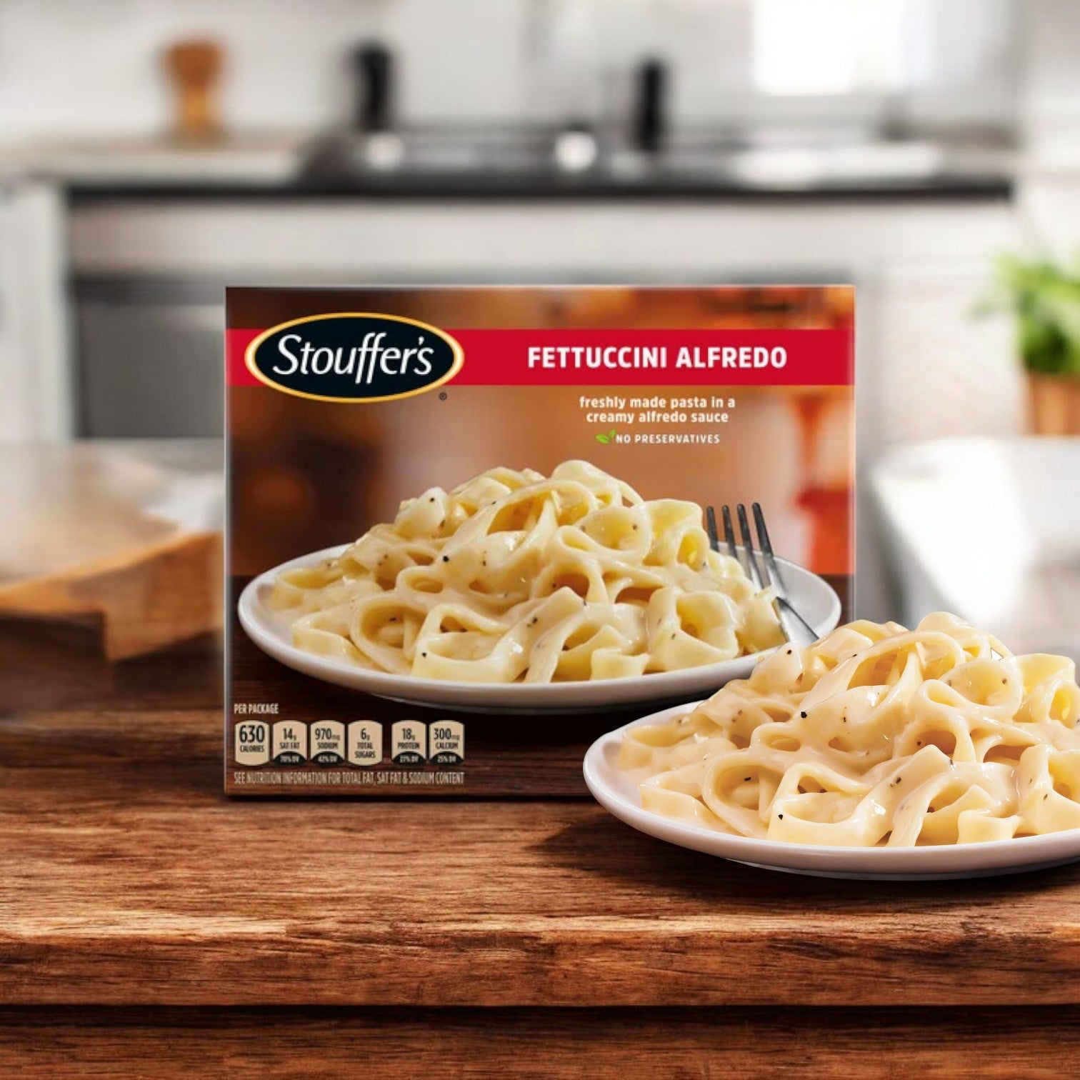 A package of Stouffer's Classics Fettuccini Alfredo is displayed on a kitchen counter beside a plate of cooked fettuccine alfredo pasta, showcasing one of the comforting meals included in the Stouffer's Bundle, which features 3 Stouffer's Classics Spaghetti With Meat Sauce, 3 Stouffer's Roast Turkey, and 3 Stouffer's Classics Fettuccini Alfredo for a total of 9 meals.