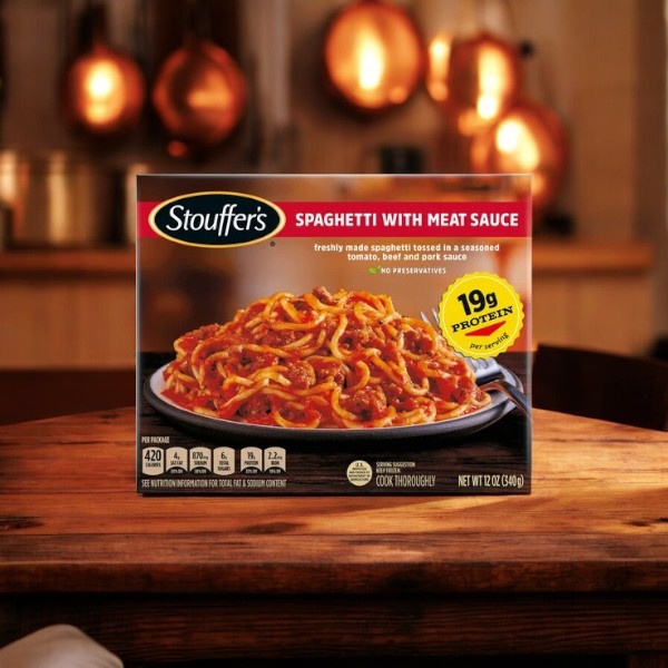 A box of Stouffer's Classics Spaghetti With Meat Sauce, 12 oz., sits on a wooden table in a kitchen, showing a plated serving of the pasta and highlighting 19g of protein per serving. Nearby, you'll spot Stouffer's Roast Turkey with creamy mashed potatoes and homestyle gravy for a comforting meal combo.