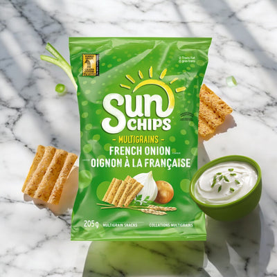 A 205g bag of Sun Chips French Onion is placed on a marble surface, next to a small green bowl of dip and chip pieces, with green onion slices scattered around. This savory snack from Sun Chips, made from whole grains, promises a delightful crunch in every bite.