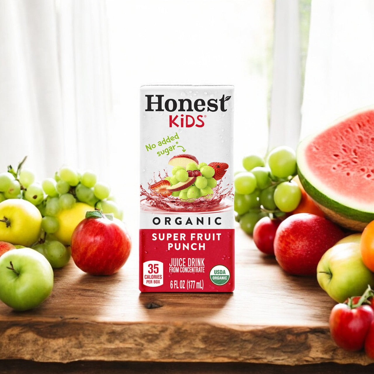 A box of Honest Kids Organic Fruit Punch (6 oz, 1 count) is placed on a wooden table surrounded by various fresh fruits, including apples, grapes, and a slice of watermelon.