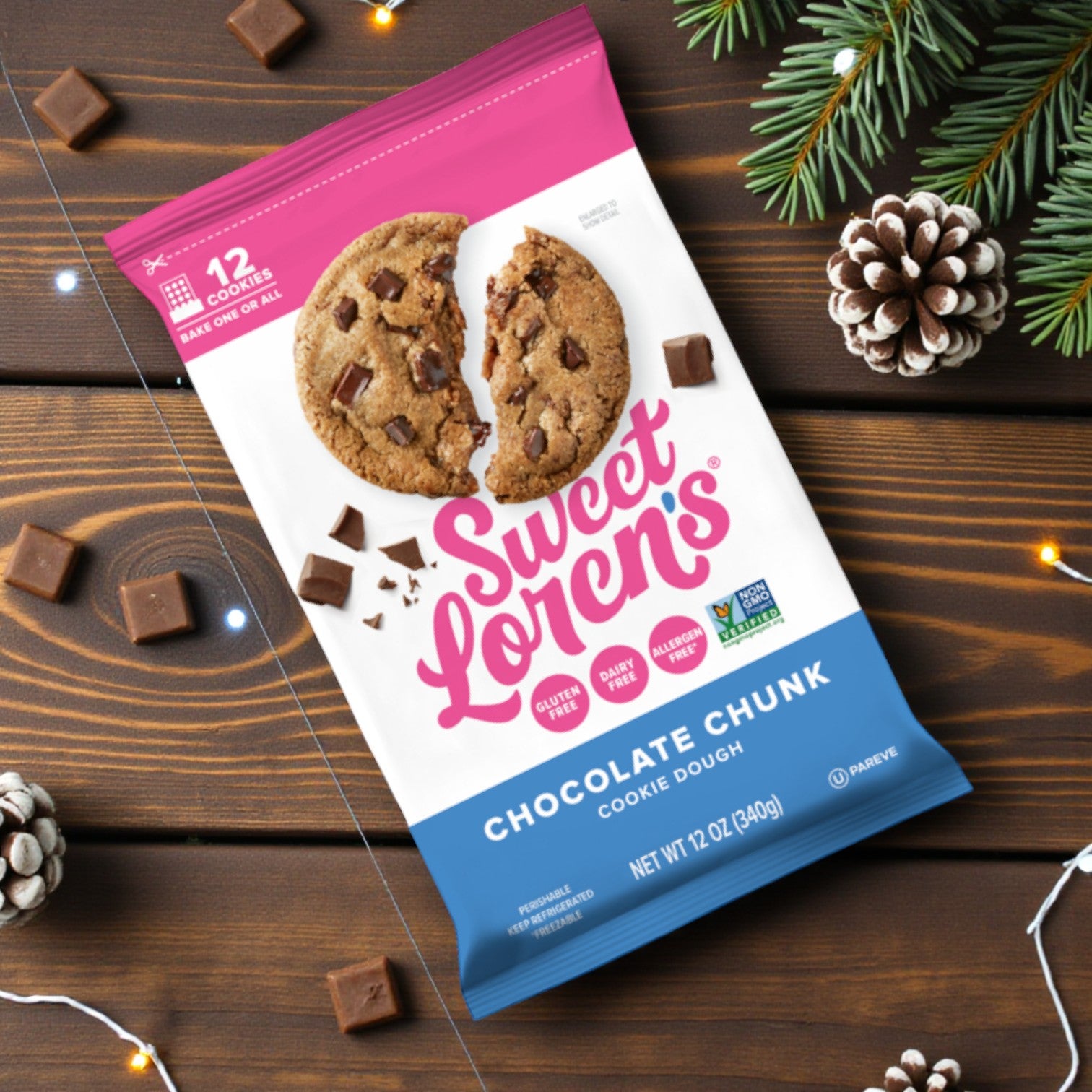 A 12 oz package of Sweet Loren’s Chocolate Chunk Cookie Dough (Gluten-Free & Vegan) sits on a wooden table with chocolate chunks, pinecones, and pine branches—ideal for anyone craving delicious vegan treats.