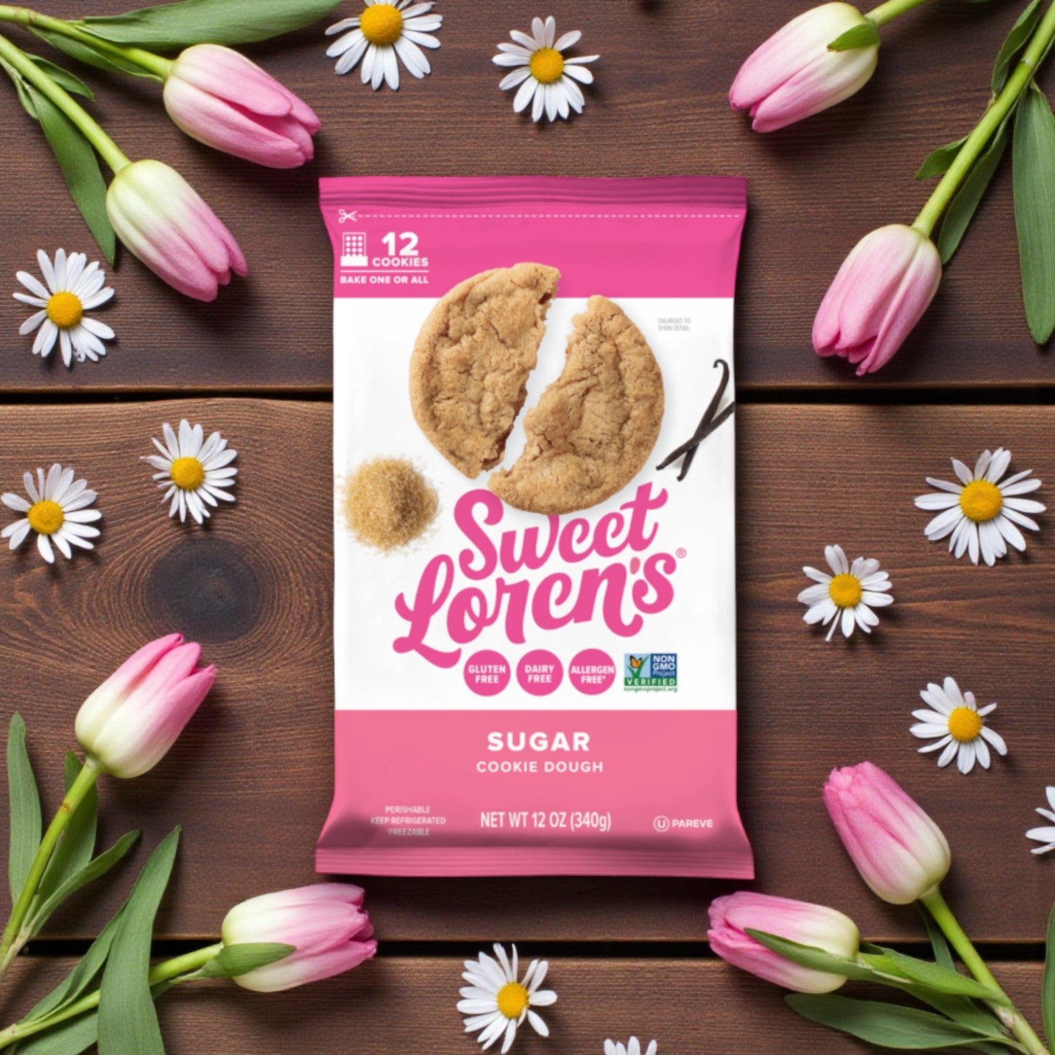 A package of Sweet Loren’s Sugar Cookie Dough – Gluten-Free & Vegan 12 oz (3-Pack) sits on a wooden surface, surrounded by pink and white tulips and daisies.