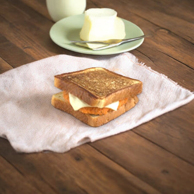 Timber Ridge Spicy Chicken & Swiss on Texas Toast is presented on a cloth napkin, accompanied by a small plate with butter and a knife, and a glass of milk in the background, all set on a wooden surface.