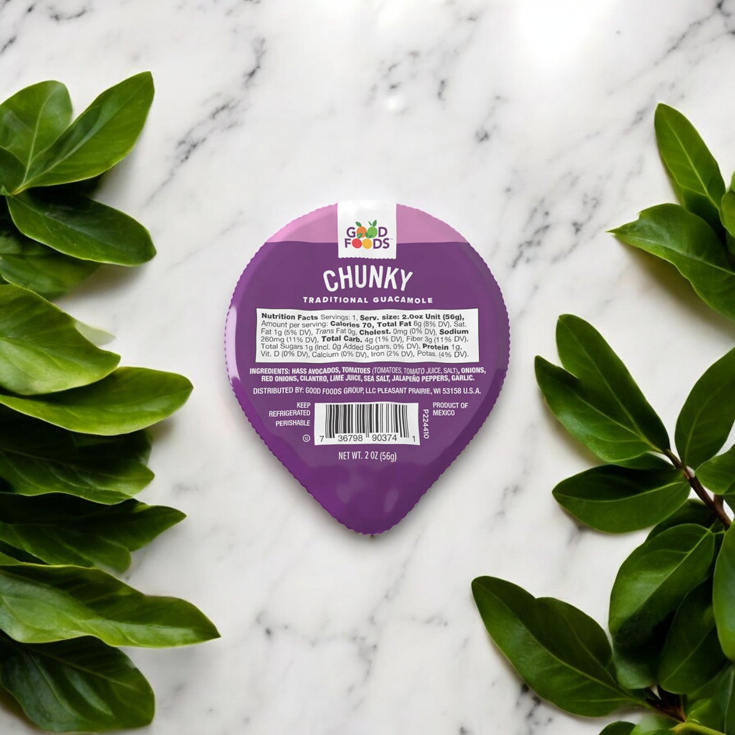 An individual serving container of Good Foods Chunky Guacamole - 1/2 oz, a healthy snack from the Good Foods brand, is placed on a marble surface, surrounded by green leaves.