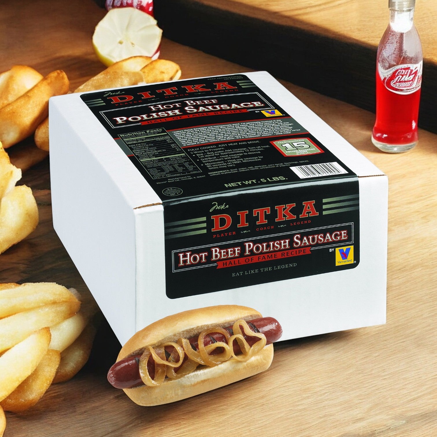 A box of Vienna Beef's Ditka Hot Beef Polish Sausage rests on a table, accompanied by a spicy sausage in a bun with mustard, crinkle-cut fries, and a glass bottle of sauce in the background.