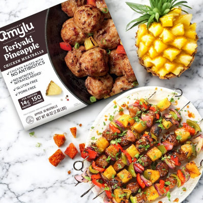 Amylu Teriyaki Pineapple Chicken Meatballs – 46 oz package showcases skewered, antibiotic-free chicken meatballs with pineapple and vibrant veggies. Enjoy diced pineapple in a half shell with red pepper pieces on a white surface.