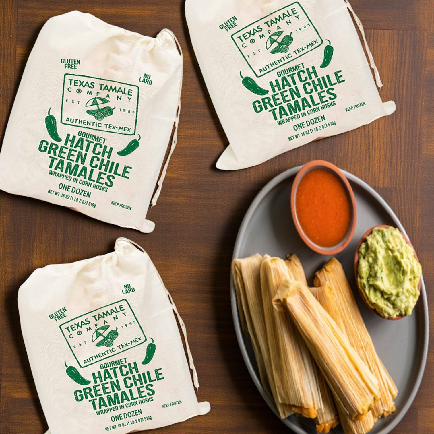 Three packs of Texas Tamale Company Hatch Green Chile Tamales – 18 oz sit beside a plate of tamales, a bowl of red sauce, and guacamole on a wooden surface.