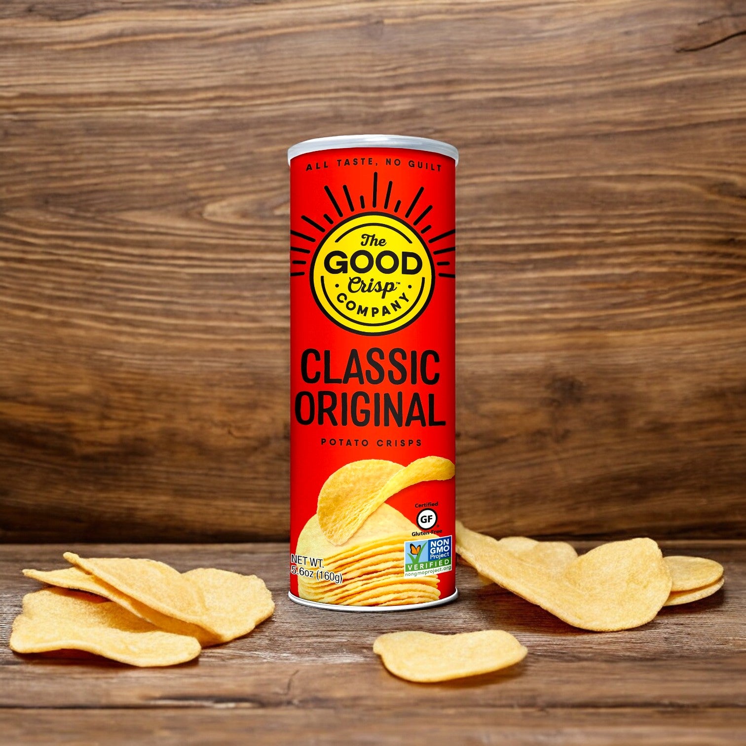 A can of The Good Crisp Company Gluten Free Original Snack Chips, 5.6 oz, is on a wooden surface, surrounded by several crisps. The packaging features a red design with yellow accents and various certification labels.