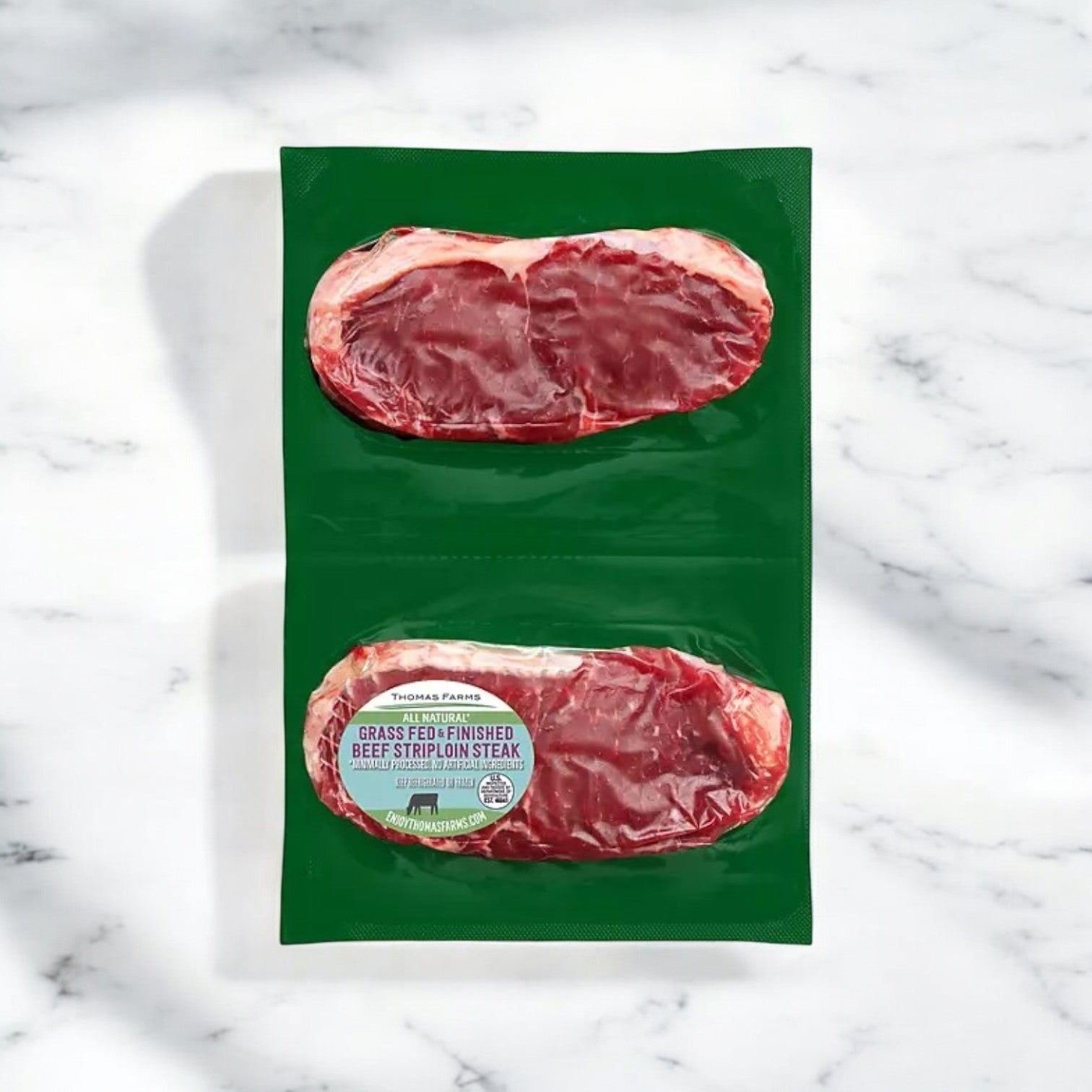 Three New York Strip Steaks, featuring premium quality grass-fed beef from Thomas Farms, are elegantly packaged in green and displayed on a marble surface.