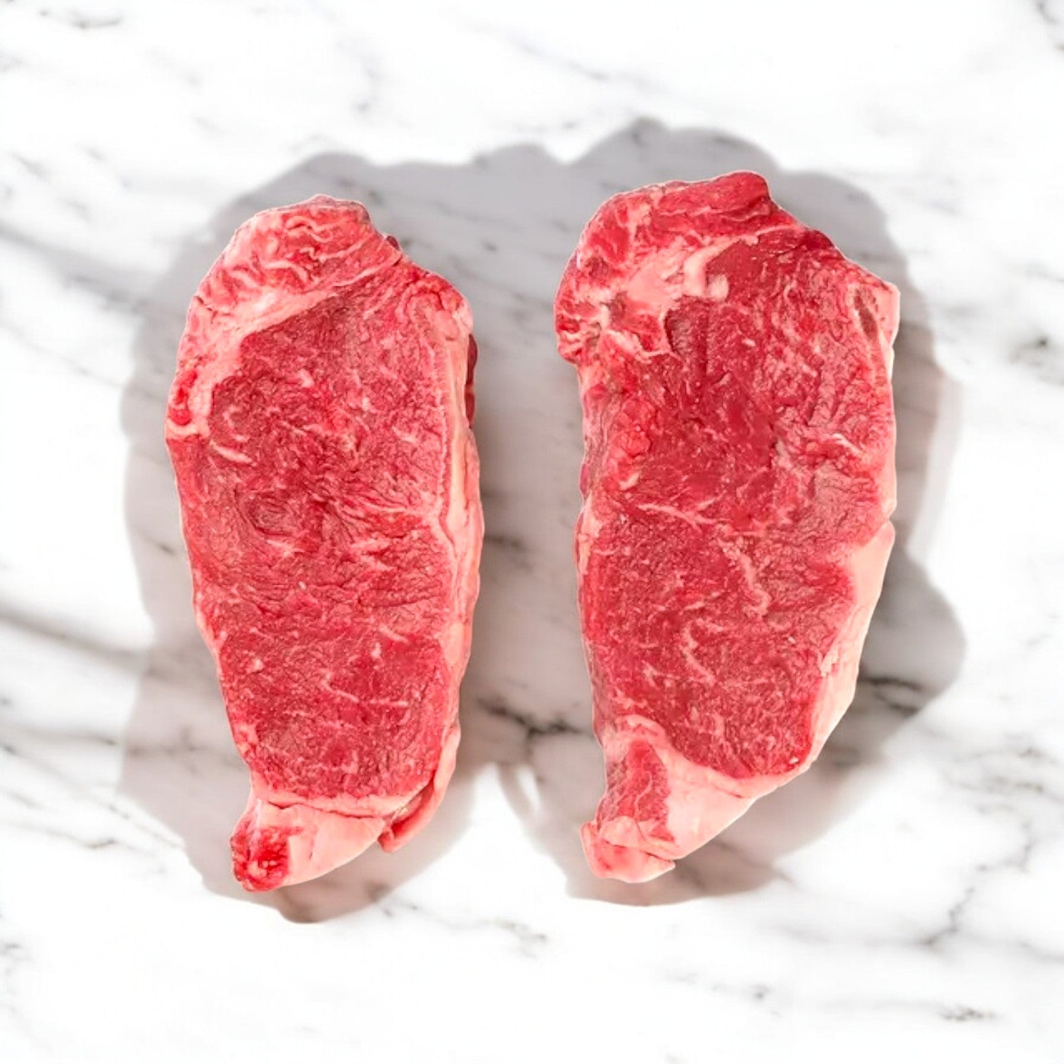 Six marbled New York Strip Steaks from Thomas Farms, showcasing premium quality, are elegantly displayed on a pristine white marble surface.