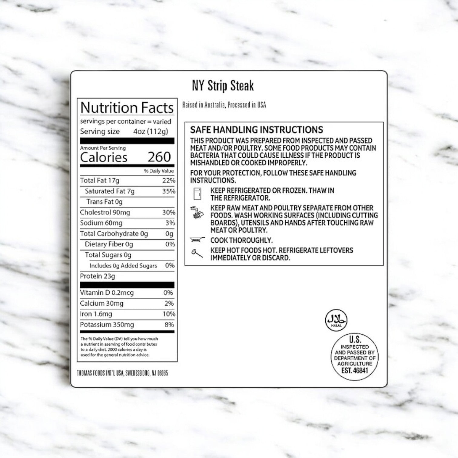 A white label on a marble background showcases the nutrition facts and safe handling instructions for Thomas Farms' New York Strip Steaks. Emphasizing its premium quality, it features storage and cooking guidelines. The USDA and Halal symbols are included, ensuring that you have the finest choice in grass-fed beef from Thomas Farms.