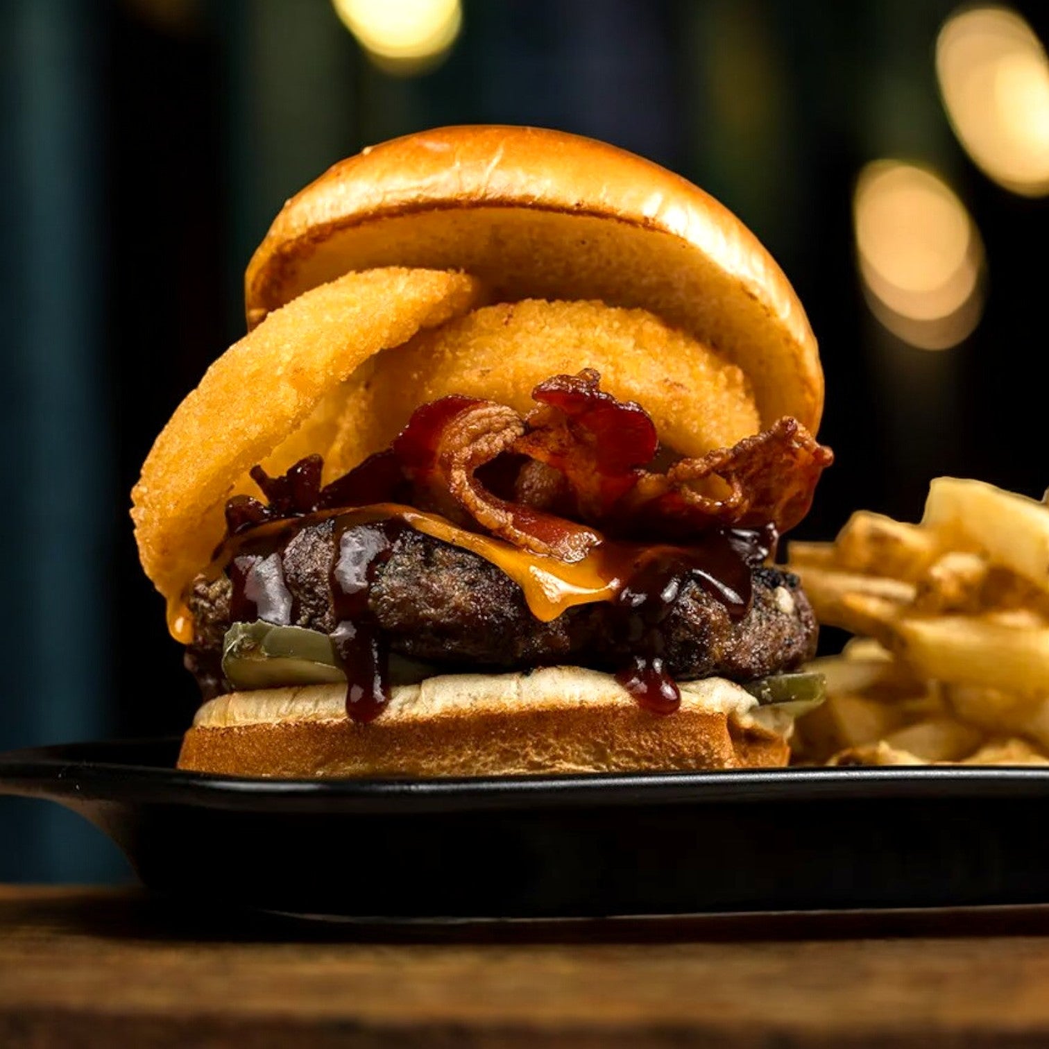 Enjoy Mino Prime Steaks' Prime Steak Burger, garnished with onion rings, bacon, cheese, and barbecue sauce on a fresh bun, with crispy fries served on a sleek black plate.