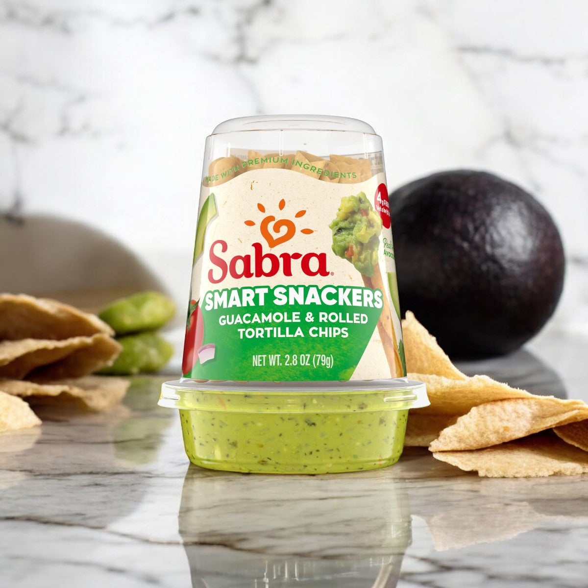 A package of Sabra Guacamole with Rolled Tortilla Chips, 2.8 oz - 1 Count, is displayed on a marble surface with an avocado and chips in the background, highlighting the use of fresh Hass avocados. This portable snack from Sabra offers a convenient and tasty option for any time of day.