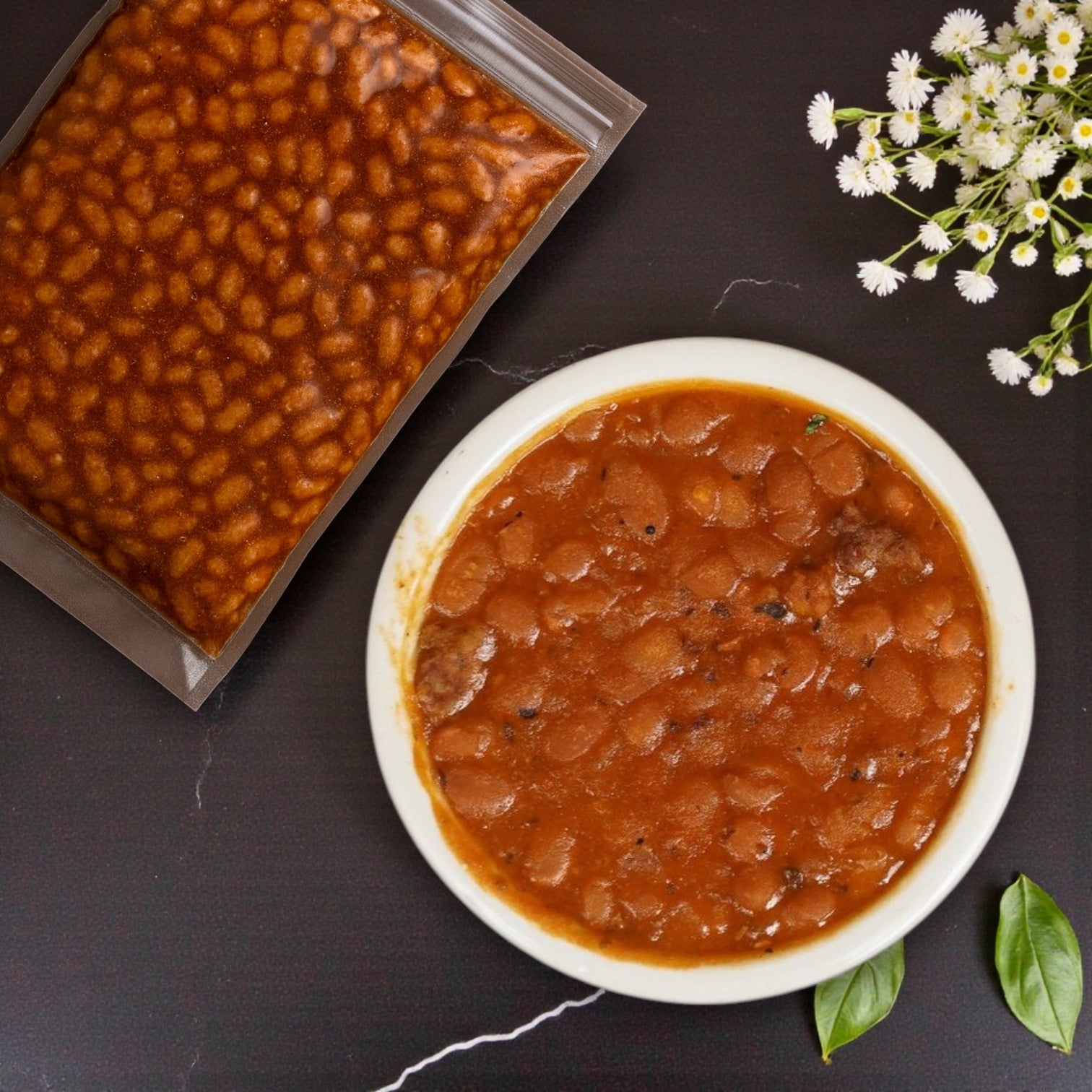 A bowl of barbecue-style baked beans in tomato sauce sits beside a sealed 6 oz TJ Ribs Baked Beans packet from TJ Ribs, accented by white flowers and green leaves on a dark surface.