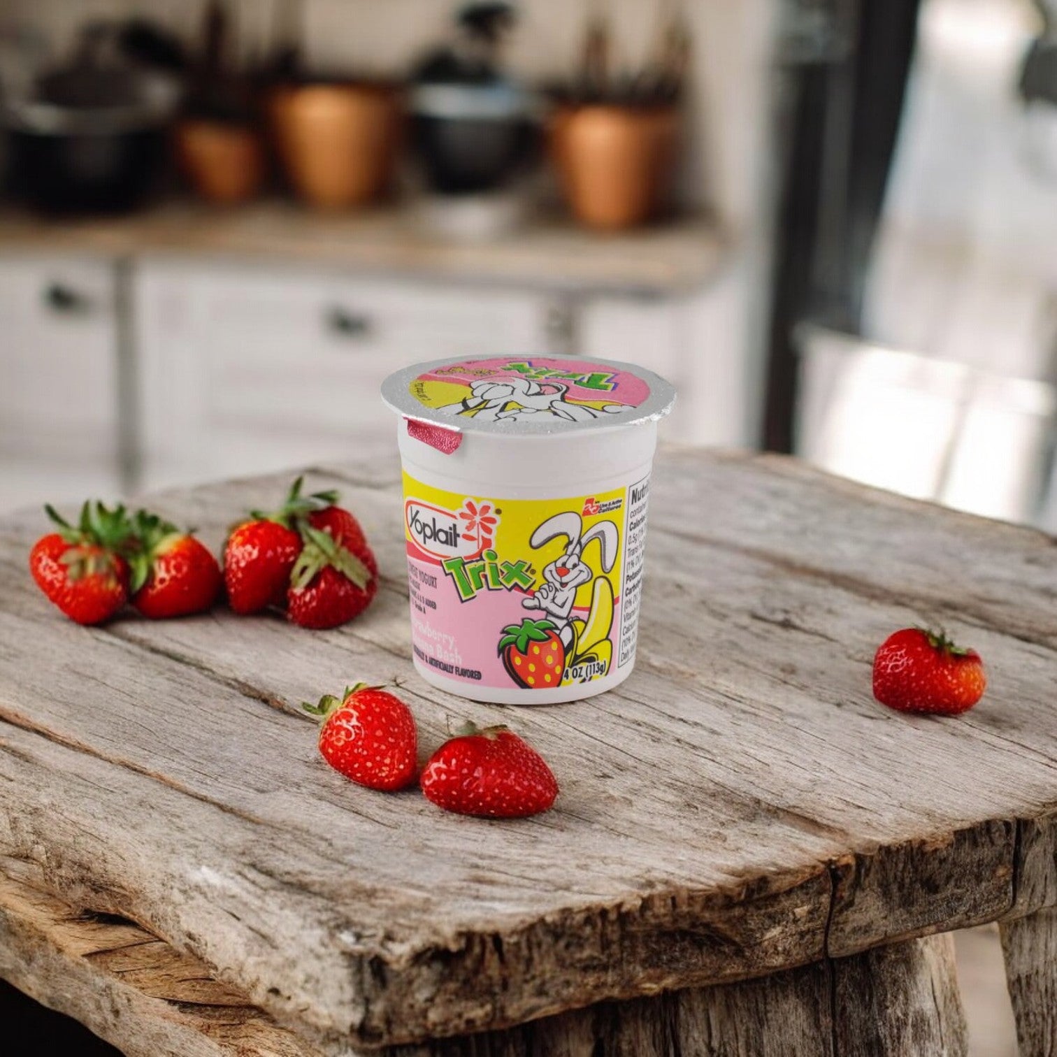 A container of Yoplait Trix Strawberry Banana Bash Yogurt, a kid-approved snack with no high fructose corn syrup, is placed on a rustic wooden table surrounded by fresh strawberries in a cozy kitchen setting.