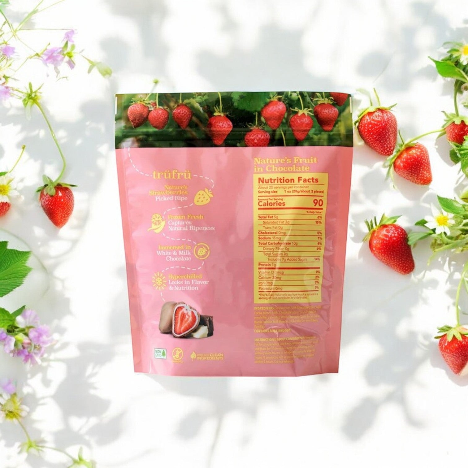 A pink Tru Fru packaging of juicy, freeze-dried strawberries covered in white and milk chocolate is placed on a light background with scattered strawberries and flowers. Nutritional information for the 20 oz. count of Tru Fru Milk Chocolate Strawberries is visible on the back.