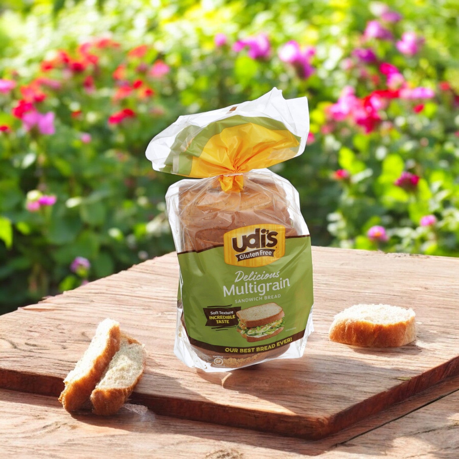 A package of Udi's Bread Loaf Multigrain Gluten Free Sliced, 24 oz., rests on a wooden table outside, accompanied by sliced pieces. Bright flowers in the background enhance the natural setting of your wholesome meal.